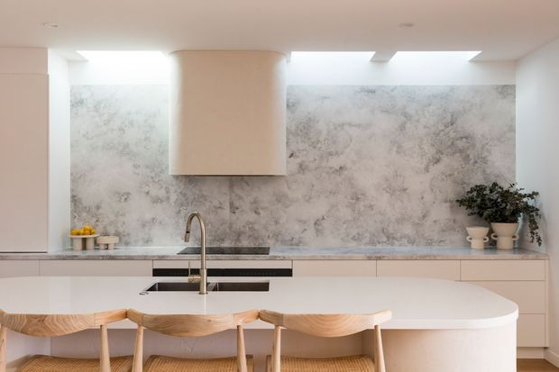 Bright contemporary kitchen with a large curved island and integrated sink, pale cabinetry, a dramatic grey-and-white marbled splashback, a sculptural rangehood, skylights, and woven timber bar stools. Potted plants and restrained styling add a warm natural accent.