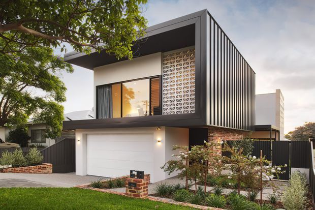 Contemporary two-storey residential house viewed from the street, featuring a strong rectangular form, projecting upper-level volume, flat roof, dark vertical battens, patterned breeze-block screening, brick detailing, and a white integrated garage door. The landscaped front garden and mature tree frame the modern facade.