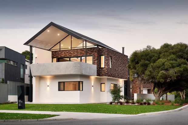Contemporary two-storey residential house photographed from the street, featuring a prominent pitched gable roof with exposed triangular glazing, white rendered walls, a curved concrete balcony, dark metal cladding on the upper side volume, and patterned dark brickwork. The home is set within a landscaped suburban frontage.
