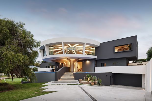 Exterior view of a contemporary multi-level residence with a dramatic curved upper-storey balcony and white sculptural roof form, dark charcoal rendered walls, extensive glazing, a broad entry staircase, landscaped front garden, and an integrated double garage. The composition highlights the home's modern facade and flat-roofed geometric forms.