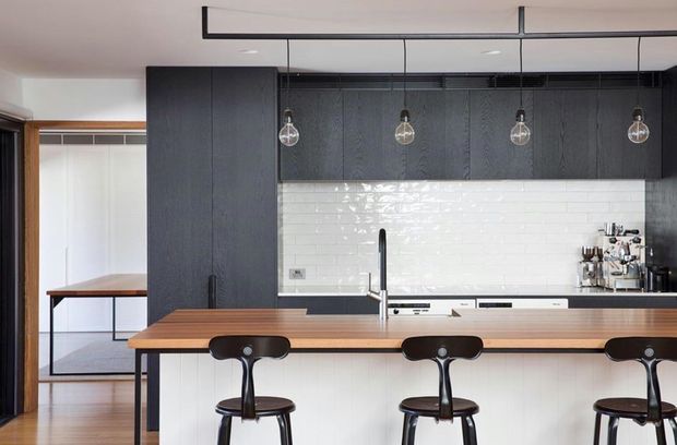 Contemporary kitchen with a long timber island and three black bar stools, dark charcoal overhead cabinetry, a glossy white subway-tile splashback, black-framed pendant lighting, and a stainless-steel espresso machine. An adjacent dining or workspace area is visible through the opening at left.