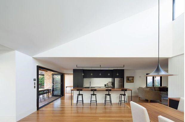 Bright open-plan contemporary kitchen and dining area with warm timber flooring, a dark kitchen island and cabinetry, four bar stools, a long dining table, and a sculptural pendant light. Large glazed openings connect the interior to an outdoor area, while a raised ceiling adds spaciousness.