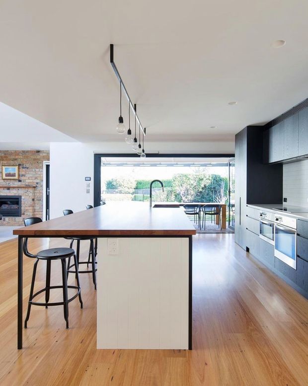 Bright open-plan kitchen with a large island, white vertical-panel cabinetry, dark blue-black full-height joinery, integrated stainless-steel appliances and a timber benchtop. The room has warm timber flooring, pendant lighting, a brick fireplace wall at left, and wide open glazing leading to an outdoor area.
