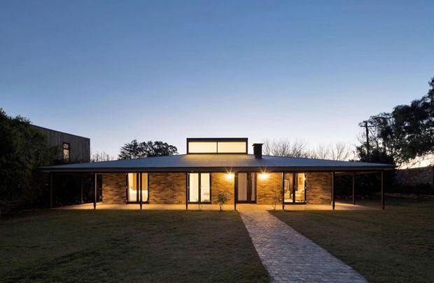 Single-storey modern brick house photographed at dusk, with a long low-pitched roof, broad covered verandah, warm-lit glazing, clerestory-like raised roof element, and a paved path leading across the lawn to the entrance.