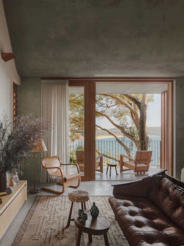 A warm, mid-century-inspired living room opening onto a waterfront balcony or terrace. The room features a dark brown leather sofa, sculptural timber lounge chair, low timber coffee tables, patterned area rug, built-in shelving and cabinetry, sheer floor-to-ceiling curtains, and a large timber-framed opening with views of water and trees. The ceiling has a mottled textured finish, and the overall composition is calm, refined, and strongly connected to the outdoors.