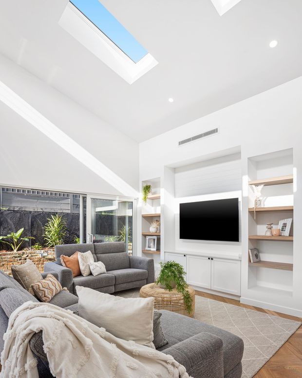 Bright contemporary living room with a large grey sectional sofa, textured cushions and throw, built-in white entertainment cabinetry and open timber shelving, a wall-mounted television, pale geometric rug, and indoor plants. Large glazed doors open to a planted outdoor area, while a skylight and recessed lighting provide abundant natural light.