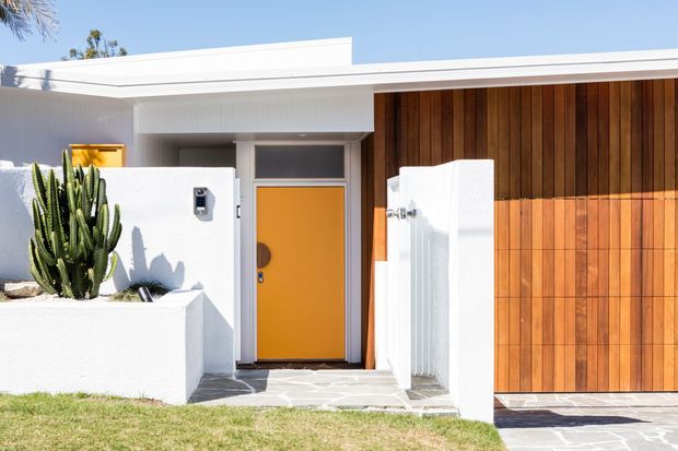 Bright contemporary single-storey home frontage with a flat roof, white rendered walls, a bold mustard-yellow entry door, vertical timber wall cladding, and a concealed garage door. A sculptural cactus planter and low white entry walls frame the paved approach.