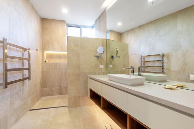 Bright contemporary bathroom featuring large beige stone-look wall and floor tiles, a walk-in shower with recessed illuminated shelving, a wall-mounted heated towel rail, a floating double vanity with white vessel basins, and a large mirror. Natural light enters through a high horizontal window.