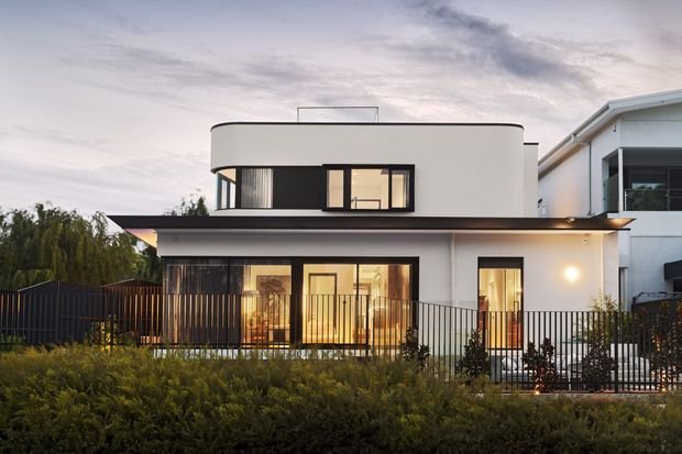 Exterior view of a contemporary two-storey residence at dusk, featuring a crisp white rendered facade, dark horizontal roof canopy, extensive black-framed glazing, a rooftop glass balustrade, and a fenced landscaped front garden.