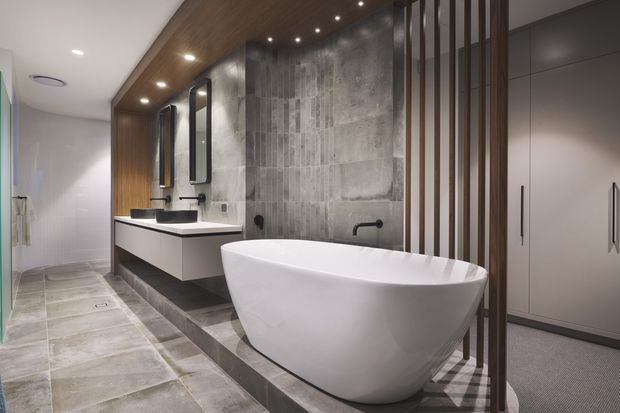 Contemporary bathroom featuring a freestanding white bathtub, double floating vanity with black basins, large-format grey stone-look floor tiles, a textured grey feature wall, dark timber ceiling and cabinetry, and a vertical timber privacy screen. A carpeted wardrobe or adjoining room is visible beyond the screen.