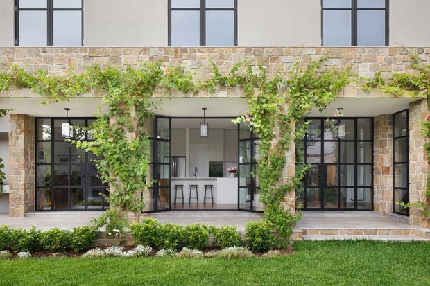 Exterior view of a two-storey residence with a stone-clad facade, dark metal-framed windows and glazed doors, and a covered ground-floor terrace. Climbing greenery is trained around the structural openings, with a visible contemporary kitchen behind the central glazing and a manicured lawn and hedge in the foreground.