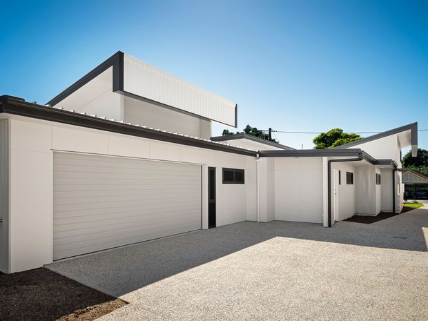 A bright exterior view of a contemporary multi-level residential building, dominated by white rendered walls, dark trim, flat roof forms and a large integrated garage door. The broad light-coloured paved forecourt and covered entry walkway are clearly visible, with a small portion of greenery in the background.