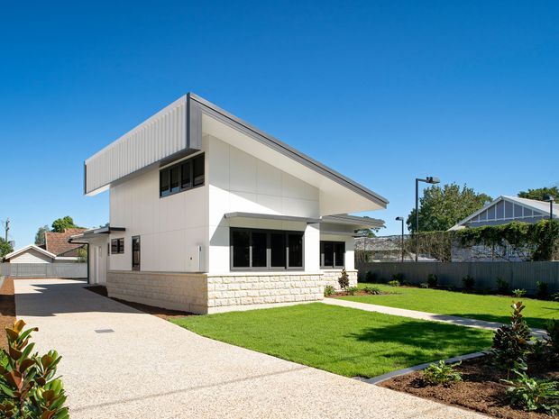 A bright, contemporary single-storey house viewed from the front, featuring a sharply angled skillion-style roof, white panelled exterior walls, dark-framed windows, and a pale stone masonry base. The property has a broad textured concrete driveway, neatly landscaped lawn and garden beds, with neighbouring homes visible beyond the boundary fence under a clear blue sky.