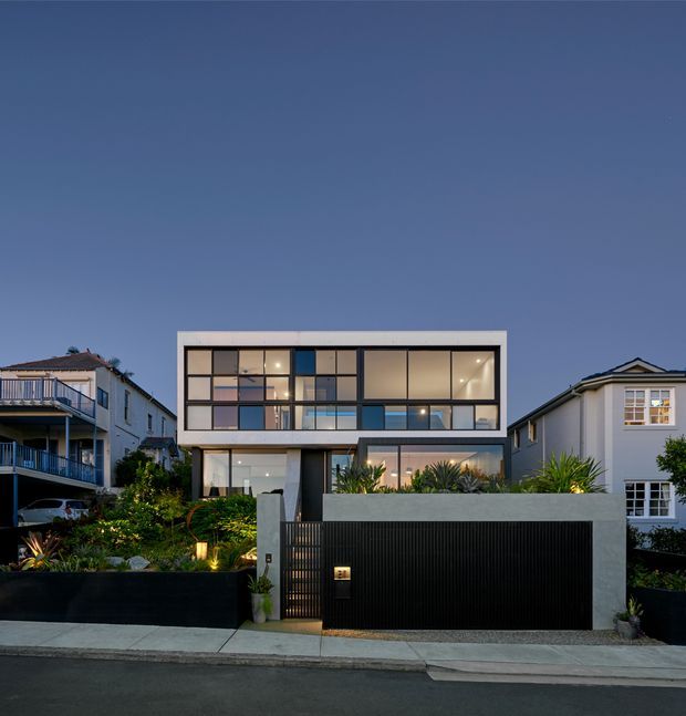 Dusk exterior view of a contemporary multi-storey urban residence. The home features a rectilinear white-framed facade, extensive floor-to-ceiling glazing, a distinctive grid of upper-level windows with varied opaque and transparent panels, a dark vertical-slat garage or entry gate, and landscaped planting behind a low front wall. Neighbouring residential buildings and the street frontage are visible.