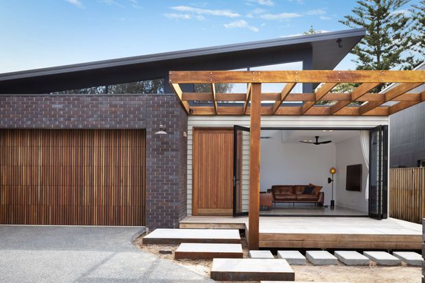 Contemporary single-storey residential exterior with dark brickwork, vertical timber garage and entry doors, and a timber pergola framing a wide open living area. The recessed interior includes a sofa, wall-mounted television, ceiling fan, and polished light floor, while stepped concrete pavers lead to the entrance. A low-pitched projecting roof and clean geometric lines define the facade.