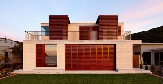 Front exterior view of a contemporary two-storey residence with a pale masonry facade, extensive dark timber louvre screens, a flat roof, upper-level terrace railings, and broad glazed openings. The house sits in a suburban setting with a landscaped lawn and neighbouring buildings visible at the edges.
