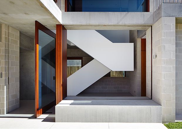 A modern exterior entry sequence featuring a recessed doorway with a warm timber-framed glass door, pale concrete slab construction, light grey block walls, and a bold sculptural white staircase visible within a covered double-height void. Strong geometric forms and contrasting timber, concrete, and masonry create a minimalist architectural composition.
