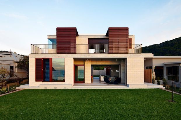 A contemporary two-storey residential house viewed from the front across a broad lawn. The façade combines pale masonry cladding with strong rectangular forms, dark red-brown timber or louvred privacy screens, expansive glazing, and slim metal balcony railings. A recessed upper-level terrace and covered ground-floor outdoor living area are visible, with neighbouring homes and a vegetated hillside in the background.