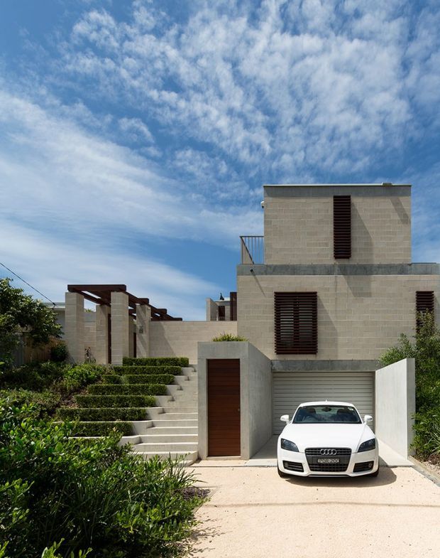 Exterior view of a contemporary multi-level residence with pale masonry walls, projecting cubic forms, dark timber louvres, a flat roof, and an integrated garage. A stepped landscaped garden and broad concrete driveway lead to the entry and parked car. The image is photographed under a bright blue sky.