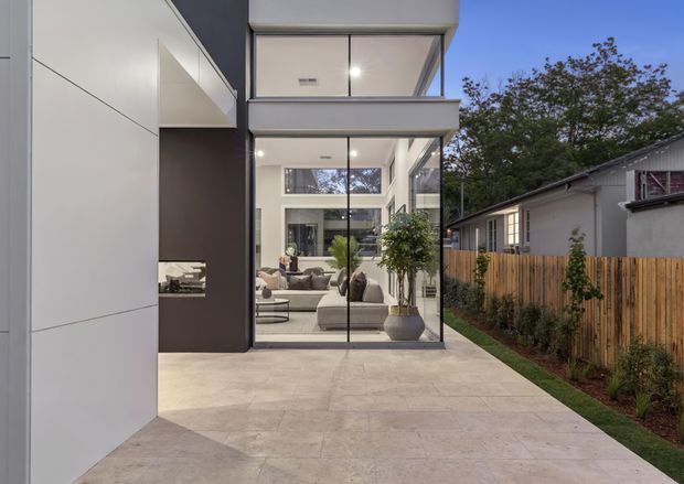 Exterior view of a contemporary multi-level residential extension with a tall glazed living area opening onto a broad pale paved terrace. The facade combines large frameless-looking glass panels, crisp white wall planes, and a dark vertical feature wall. Through the glazing, a bright living room with sofas, a coffee table, plants, and a high clerestory window is visible. A timber boundary fence, neighbouring low-rise dwelling, and landscaped planting frame the suburban setting.
