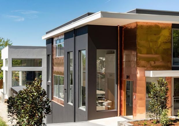 Exterior view of a contemporary multi-level residence with a flat roof, strong geometric volumes, dark vertical cladding, extensive glazing, and a prominent copper-toned metal facade panel. Landscaped shrubs and a paved entry are visible in the foreground.
