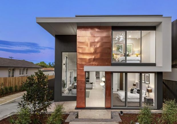 Front exterior view of a contemporary multi-storey residence with a strong rectangular composition, broad flat roof overhangs, extensive floor-to-ceiling glazing, dark framing, and a central copper-toned feature wall. Interior living and bedroom spaces are visible through the windows, with a landscaped front path and neighbouring homes around the property.