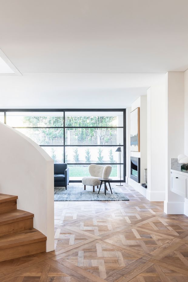Bright contemporary open-plan living area viewed from a timber staircase. The space features expansive black-framed floor-to-ceiling glazing, a sculptural lounge chair, dark sofa, fireplace and built-in white cabinetry. The floor is finished in an elaborate geometric parquet pattern, with white walls and ceiling creating an airy, refined interior.