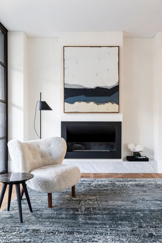 A bright, refined living room centred on a dark built-in fireplace with a pale stone hearth and abstract artwork above. The space features white walls, tall black-framed glazing, a sculptural cream boucle armchair, a small dark side table, a black floor lamp, a low decorative object, and a large distressed grey-blue area rug over timber flooring.