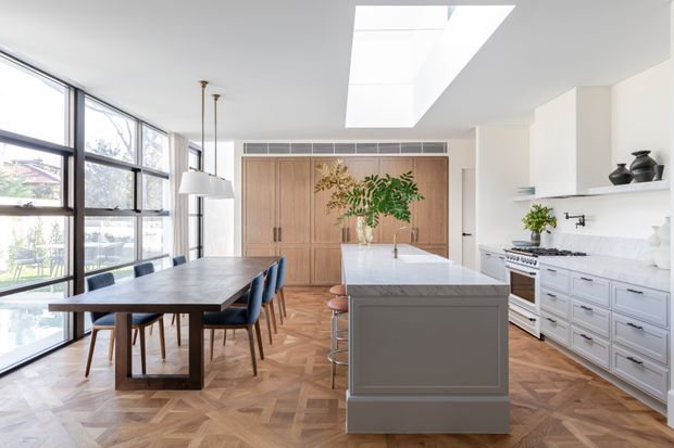 Bright, refined open-plan kitchen and dining area with expansive black-framed windows, a large skylight, herringbone timber flooring, white shaker-style cabinetry, a marble-look island and splashback, and a built-in timber feature wall with concealed storage. A substantial timber dining table and upholstered chairs sit beside the island, creating an airy, polished contemporary interior.