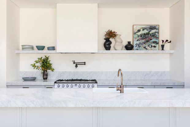 Bright, refined contemporary kitchen featuring a large white marble-look island benchtop, pale cabinetry, a matching splashback, a freestanding stainless-steel range, and a brushed brass tap. Open shelving displays ceramics, vases, artwork, and greenery.