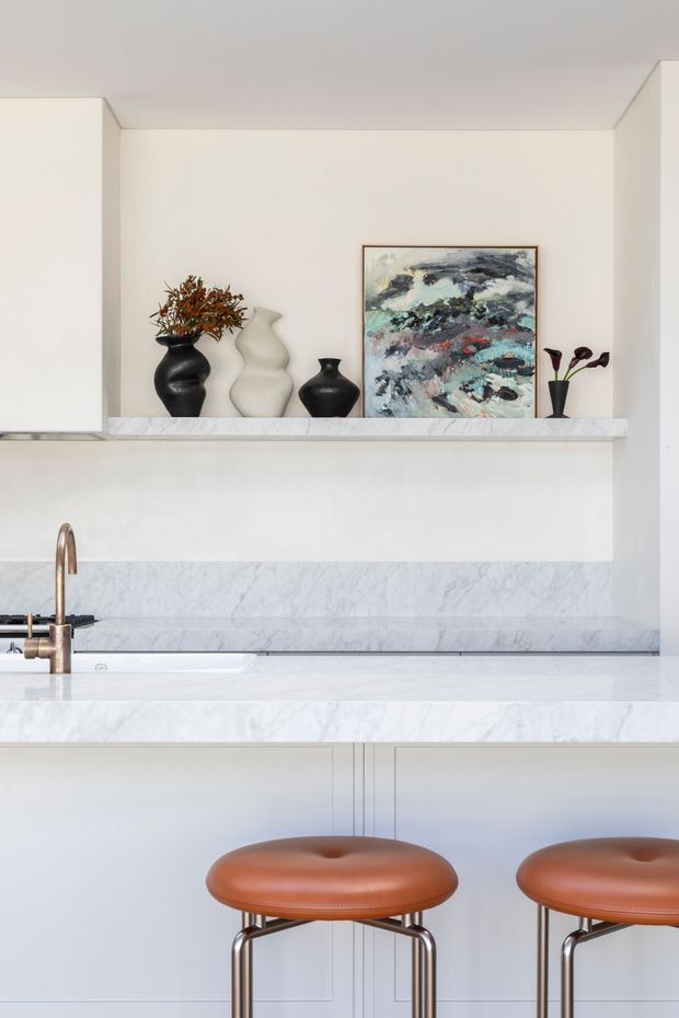 A refined contemporary kitchen with white cabinetry and walls, a pale veined marble benchtop and backsplash, and a bronze-toned tap. A raised open shelf displays sculptural black and white ceramic vessels, dried foliage, and framed abstract artwork. Two tan leather counter stools sit at the island, adding warmth to the otherwise light, minimal space.