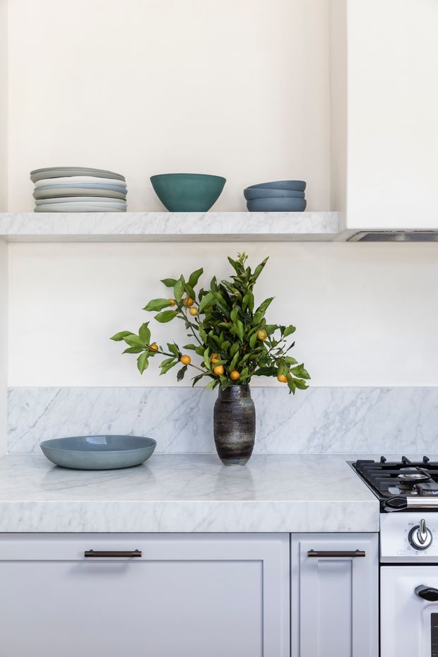 A close-up view of a refined kitchen with white cabinetry, a white and grey-veined marble benchtop and splashback, dark metal hardware, and a gas cooktop at right. Open shelving displays stacked ceramic plates and bowls, while a dark vase with a leafy citrus branch provides a natural green accent.