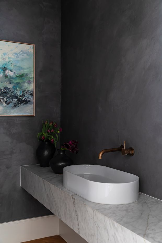 Moody contemporary bathroom featuring a white oval vessel basin set on a projecting marble vanity, dark textured plaster walls, and a wall-mounted aged-bronze tap. Black decorative vases with foliage and framed artwork add contrast, while timber flooring is visible below.