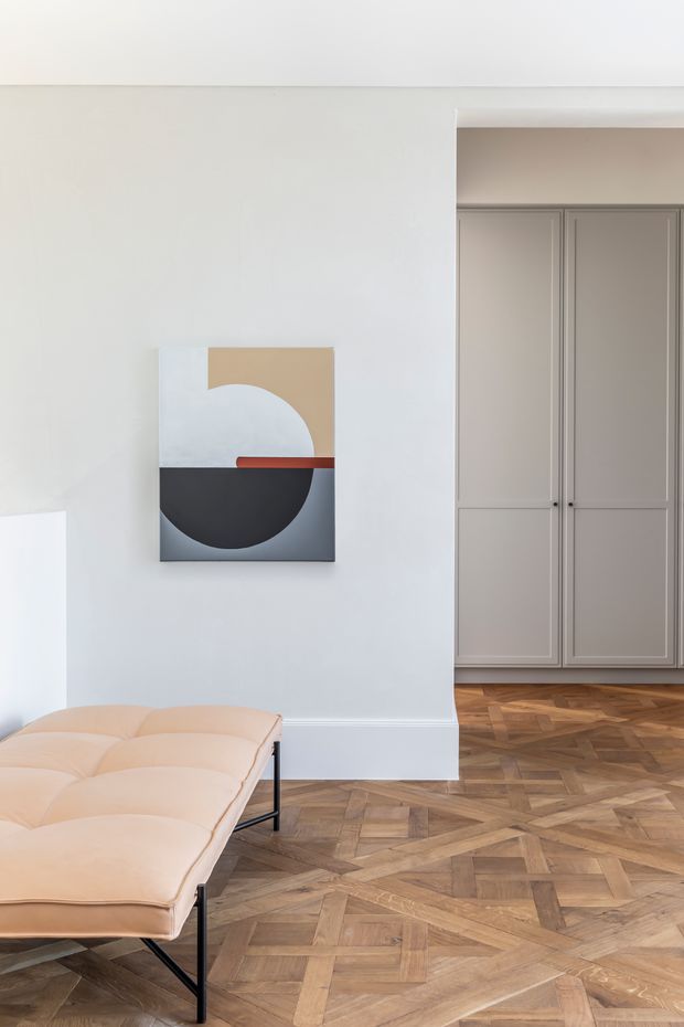 Bright, minimalist interior with a pale upholstered bench in the foreground, a geometric abstract artwork on a white wall, and a taupe built-in wardrobe or cabinetry visible through an opening. The room features an intricate warm timber parquet floor and clean white architectural detailing.
