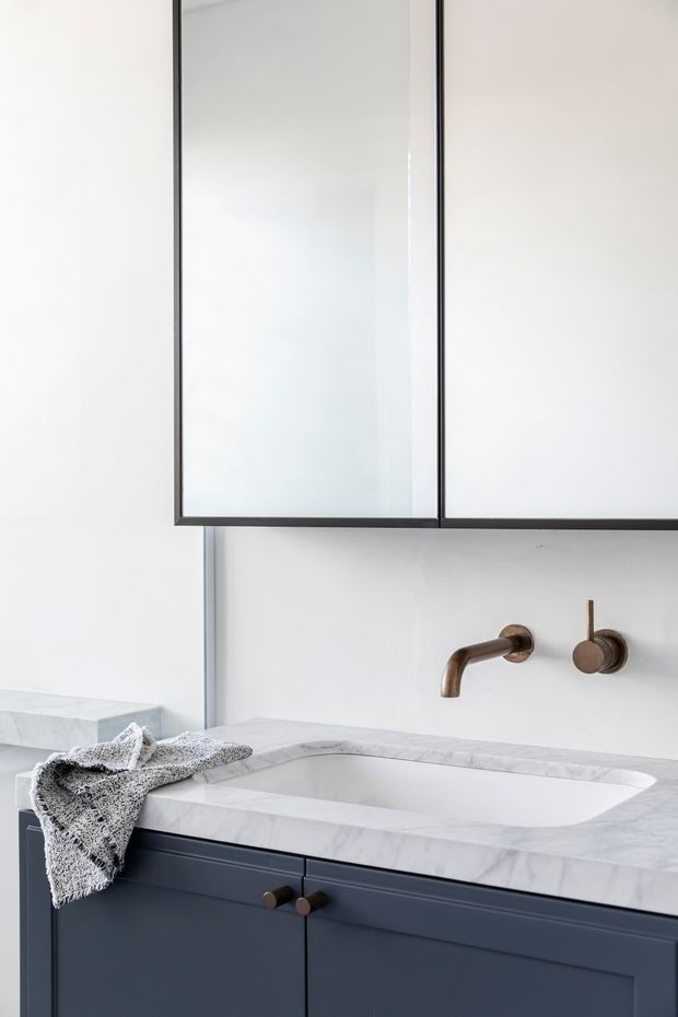Close-up of a refined bathroom vanity with a white marble benchtop, integrated rectangular basin, dark blue-grey paneled cabinetry, wall-mounted brushed bronze tapware, and a large mirrored cabinet above. The space is bright and minimalist with subtle traditional detailing in the joinery.