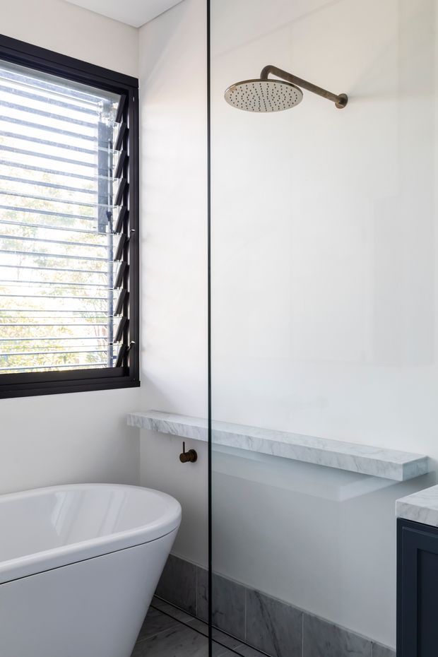 Bright contemporary bathroom with a freestanding white bathtub, frameless glass shower screen, wall-mounted rainfall shower in a bronze finish, white walls, dark-framed louvred window, grey stone or marble-look floor and skirting tiles, and a pale stone ledge with dark blue cabinetry at the edge of the frame.
