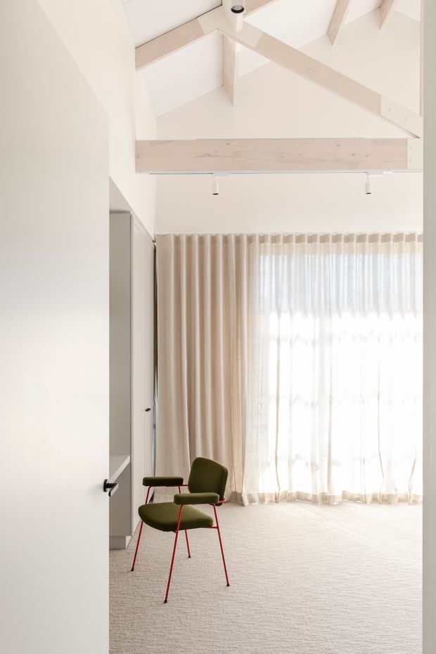 A bright, minimalist interior viewed through an open doorway. The room features pale carpet, full-height sheer and beige curtains, a large sunlit window, sparse olive-green chairs with slender red legs, white walls, and exposed pale timber roof beams creating a high vaulted ceiling.