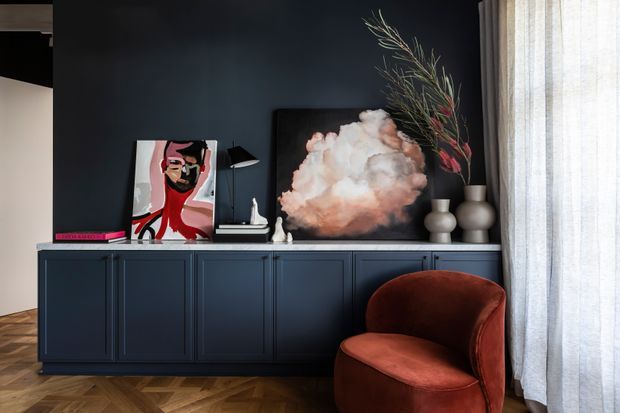 A moody, refined interior vignette featuring a dark navy built-in sideboard with panelled cabinetry, artwork, ceramic vases, and sculptural branches. A rust-red velvet armchair sits in front of the joinery beside full-height sheer curtains. The room has a warm herringbone timber floor and a dark painted wall.