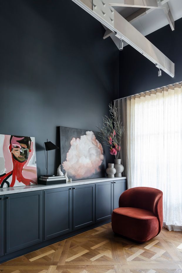 A sophisticated living room with a dark navy-blue wall and matching built-in sideboard cabinetry, herringbone timber flooring, and tall sheer curtains over a large window. The room features a sculptural rust-red lounge chair, abstract artwork, decorative vases, a black floor lamp, and exposed white ceiling beams with track lighting. The palette is moody and refined with artistic, contemporary furnishings.