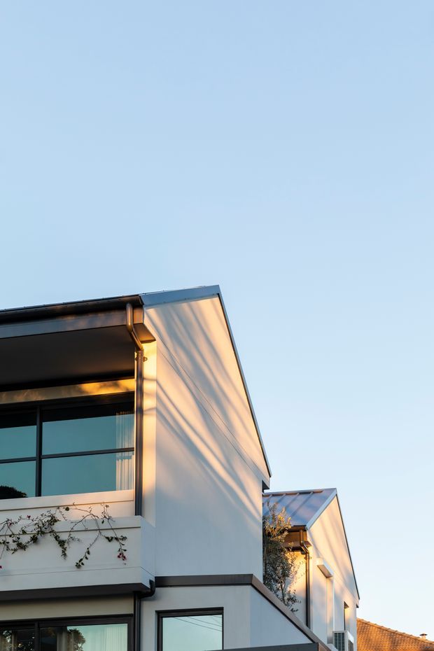 Vertical exterior architectural photograph of a contemporary multi-storey residential building, showing clean white rendered walls, dark metal-framed glazing, projecting flat roof planes and strong geometric forms. Warm low-angle sunlight creates pronounced shadows across the facade, with a small amount of climbing vegetation and neighbouring roofline visible at the edges.