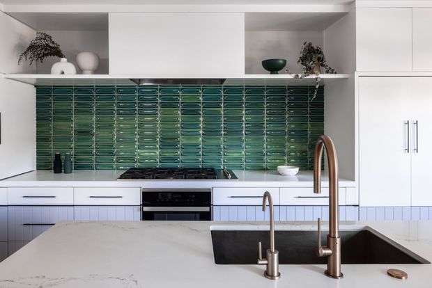 Bright contemporary kitchen featuring white built-in cabinetry, open display shelving, a glossy deep green horizontal tile splashback, dark appliances, and a large pale stone island with an inset dark sink and brushed bronze gooseneck tapware. Minimal styling includes ceramic vessels and potted greenery.