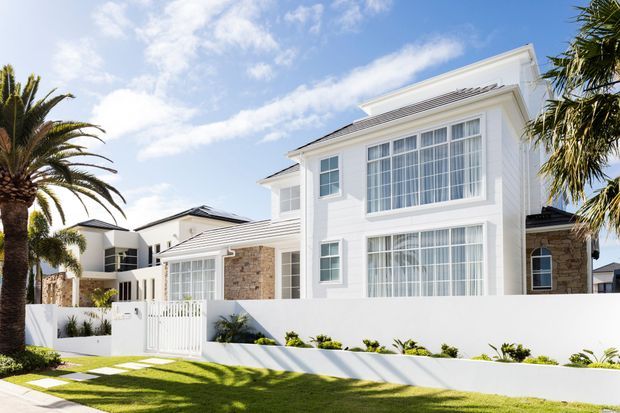 Bright exterior view of a large two-storey residential home with a white rendered facade, prominent multi-pane windows, light stone wall sections, layered low-pitched roof forms, and a white boundary wall with a gated entry. Palm trees and manicured planting frame the sunny suburban setting.