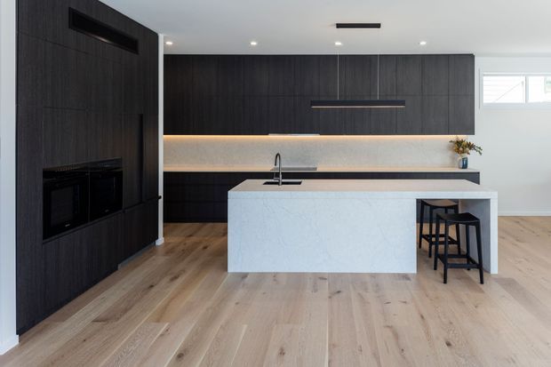 Contemporary kitchen with dark timber-look full-height cabinetry, integrated appliances, a pale stone island with undermount sink, subtle warm under-cabinet lighting, and light timber flooring. Two black stools sit at the island, with a small plant and decorative vessel on the rear benchtop.