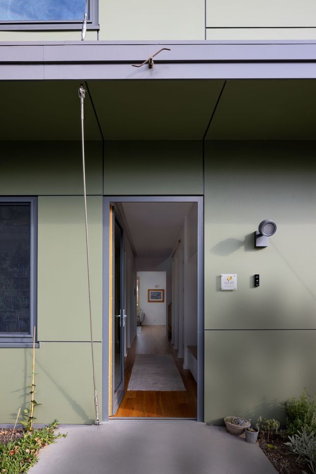 Front exterior view of a contemporary residential entry, featuring pale green panel cladding, a deep recessed canopy, dark-framed glazing, a narrow open doorway, and minimalist exterior lighting. The entry opens to a timber-floored hallway with artwork visible inside, while low planting borders the concrete approach.
