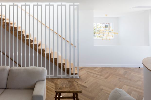 Bright contemporary living area with a warm herringbone timber floor, neutral upholstered seating, and a timber staircase screened by a distinctive white vertical metal balustrade. A sculptural modern pendant hangs near a large internal opening, with a partial curved kitchen or counter edge visible at right.