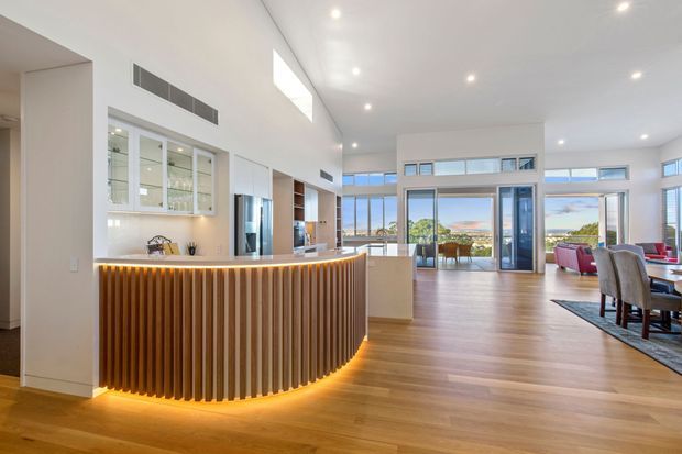 Bright open-plan contemporary interior featuring a curved kitchen island with vertical timber slat detailing and warm underlighting, white cabinetry, timber flooring, extensive glazing, and an adjoining dining and living area with views outside.