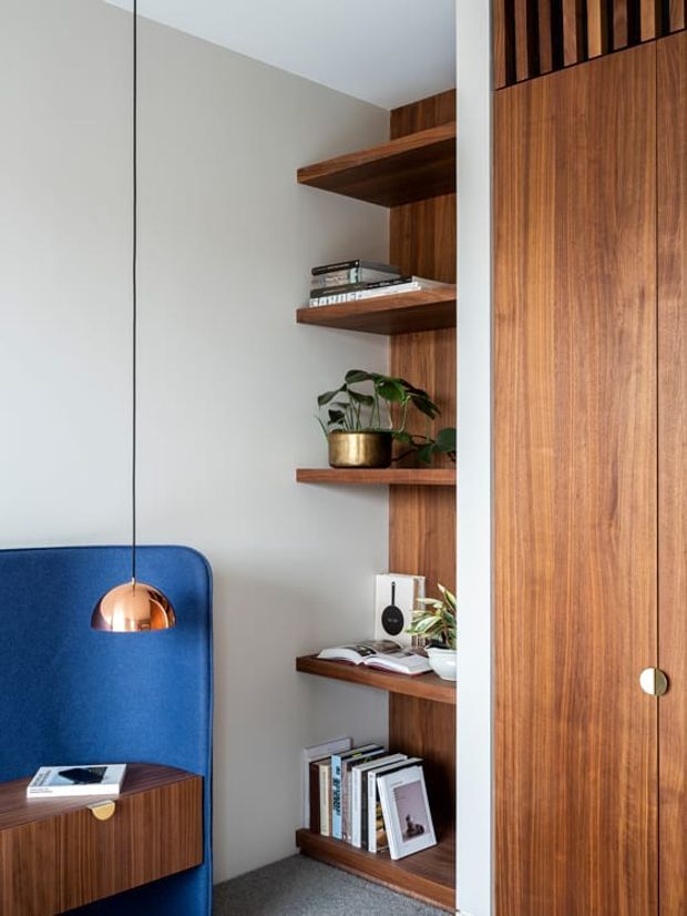 A refined built-in shelving and storage nook viewed indoors. Warm walnut-toned timber shelving and a tall matching cupboard frame books, a small plant, speaker and decor against a light grey wall. A deep blue upholstered chair and compact timber side table sit beside the joinery, with a suspended copper pendant adding a small metallic accent. The space has a clean, tailored mid-century-inspired feel.