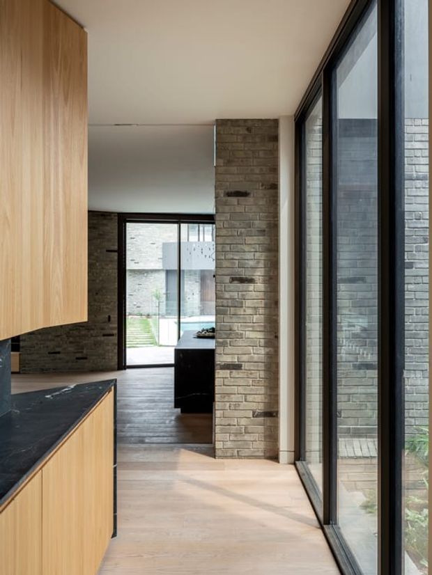 View through a contemporary kitchen toward an adjoining living area and courtyard. The space features light timber cabinetry, a dark stone benchtop, exposed brick walls and columns, pale timber flooring, and full-height black-framed glazing.