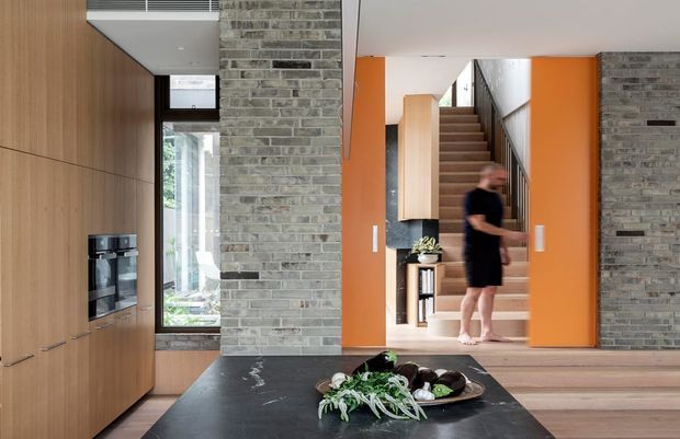 Contemporary open-plan interior showing a kitchen foreground with tall timber cabinetry, integrated appliances and a dark stone island benchtop. A central exposed grey brick wall frames a bright orange doorway leading to a timber staircase, with additional timber flooring and a glazed opening bringing in natural light.