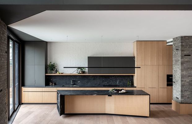 A contemporary kitchen with a large central island, light timber cabinetry, dark stone benchtops and splashback, integrated appliances, open shelving, and a brick-clad structural wall. Full-height glazing and pale timber flooring bring natural light into the space.